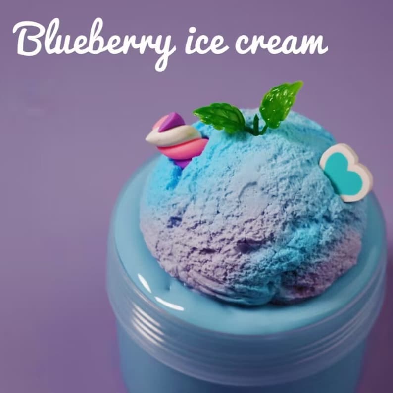 May include: A scoop of blueberry ice cream in a light blue container. The ice cream is a blend of blue and purple hues, topped with a decorative swirl, green leaves, and a blue and white butterfly accent. The text "Blueberry ice cream" is at the top.