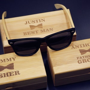 May include: Three wooden boxes with engraved text and a bow tie design. The boxes are labeled "Justin Best Man", "Sammy Usher", and "Anthony Father of the Groom". A pair of black sunglasses with wooden arms is on top of the "Justin Best Man" box.