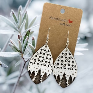 May include: A pair of wooden earrings with a white background and a black silhouette of a forest. The earrings are shaped like teardrops and have a delicate, intricate design. The earrings are hanging from silver hooks and have a "Handmade with love" tag attached.