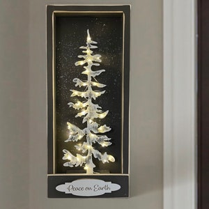 May include: A black framed wall decor with a white silhouette of a pine tree with lights inside. The frame has a white oval with the text "Peace on Earth" in black.
