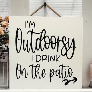May include: White wooden sign with black text that reads "I'm Outdoorsy I Drink On The Patio" with a black arrow pointing to the right.