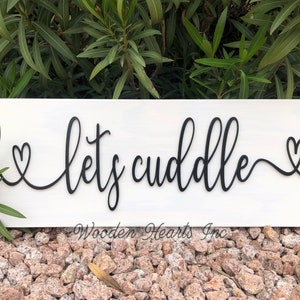 May include: White wooden sign with black lettering that reads "let's cuddle" with a heart on each side of the text.