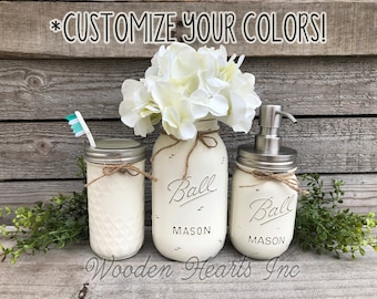 May include: Three white painted mason jars with black lettering that reads "Ball Mason" and a brown twine bow. The jars are used as bathroom decor, with one holding a toothbrush, one holding soap, and one holding flowers.