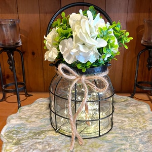 May include: A decorative glass jar with a black metal cage and handle, filled with white flowers and greenery. The jar is tied with twine and contains fairy lights. Two black metal candle holders flank the jar.