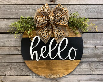 May include: A round wooden door hanger with a black and white striped design. The word "hello" is written in white script on a black background. The door hanger is decorated with a leopard print bow and green foliage.