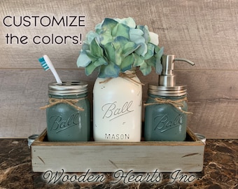 May include: A set of three mason jars painted in a teal green color with the word "Ball" printed on each jar. The jars are arranged on a wooden tray with a white background. The jars are used for bathroom storage and include a toothbrush holder, soap dispenser, and a jar for cotton balls or swabs. The text "Customize the colors!" is displayed at the top of the image. The text "Wooden Hearts Inc" is displayed at the bottom of the image.