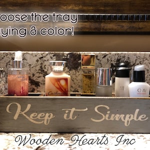 May include: A gray wooden tray with the words "Keep it Simple" in white lettering. The tray is sitting on a countertop and has several bottles of lotion and perfume on it. The tray is made by Wooden Hearts Inc.