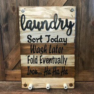 May include: A rustic wooden sign with the word "laundry" in black script lettering. The sign has four lines of text below the word "laundry" that read "Sort Today", "Wash Later", "Fold Eventually", and "Iron... Ha Ha Ha". The sign has four white ceramic hooks at the bottom.