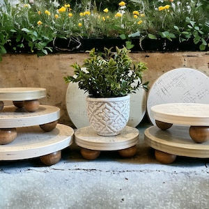 May include: Set of four white wooden tiered stands with distressed paint and round wooden legs. The stands are stacked in a pyramid shape with a potted plant on the top stand.