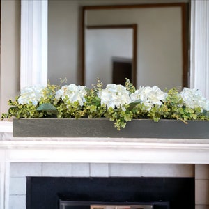 May include: A long, rectangular wooden planter box with a dark gray finish. The planter is filled with white hydrangea flowers and green foliage. The planter is sitting on a white mantel above a fireplace.