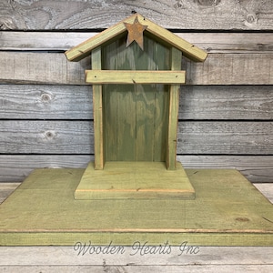 May include: A green wooden nativity scene with a star on top. The scene is on a wooden base and has a simple design. The text "Wooden Hearts Inc." is visible at the bottom of the image.