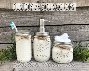 May include: Set of three white painted mason jars with silver lids and twine accents. The middle jar has a soap dispenser pump and the text "Ball Mason" printed on the front. The jar on the left has a toothbrush inside and the jar on the right has cotton balls inside. The jars are on a wooden background.