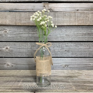 May include: A clear glass vase with a burlap ribbon tied around the neck, filled with a bouquet of white baby's breath flowers. The vase is sitting on a wooden surface.