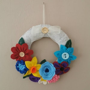 Pode incluir: Uma grinalda de flores de feltro feita à mão com uma variedade de flores coloridas, incluindo vermelhas, amarelas, azuis e roxas. A grinalda é adornada com um laço branco e um botão de madeira. Para decoração de parede.