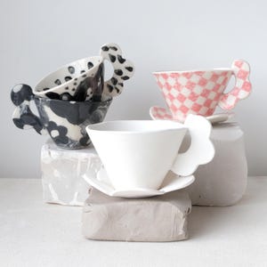Può includere: Quattro tazze da tè in ceramica con design unici. Una è bianca, un'altra ha un motivo a scacchi rosa e bianco, e due presentano motivi floreali e maculati in bianco e nero. Ogni tazza ha un manico distintivo a forma di fiore.