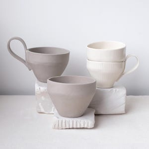 Peut inclure: Ensemble de tasses et de bols en céramique aux tons neutres. Les tasses ont des anses, et les bols présentent un design simple et moderne. Les pièces sont disposées sur des blocs blancs texturés, créant une esthétique minimaliste. La palette de couleurs est douce et discrète.