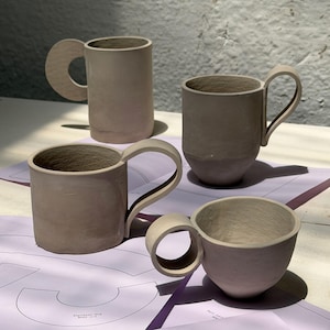 May include: Four handmade ceramic mugs in varying shapes and sizes. The mugs are a muted, earthy tone, with unique handle designs. The mugs are sitting on a purple surface with faint pattern outlines. The mugs are likely for coffee or tea.