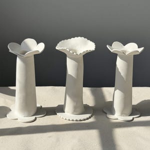 May include: Three white ceramic vases with a floral design. Each vase has a tall, cylindrical body and a flower-shaped top. The vases are arranged on a neutral surface, with soft shadows adding depth.