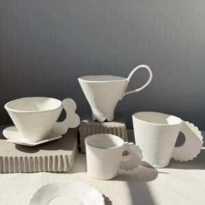 May include: A selection of white ceramic teacups and saucers. The cups have distinctive handle designs, including a petal-shaped handle and a loop handle. Some cups are on a square saucer. The cups are displayed on a neutral surface.