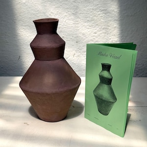 May include: A brown ceramic vase with a geometric design. The vase has a wide base, a narrow neck, and a series of stepped sections. Next to the vase is a green booklet with the title "Madre Vessel" and a drawing of the vase.