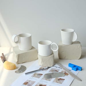 May include: Three white ceramic mugs with rounded handles sit on beige clay blocks. Pottery tools, including a sponge, a knife, and a blue plastic tool, are arranged on a white surface. A sheet with illustrated instructions is also present.