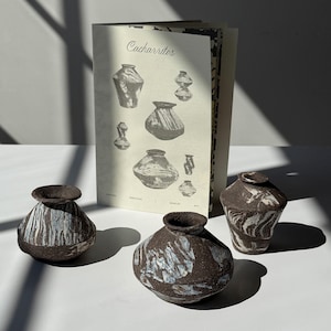 May include: Three small, dark brown ceramic vases with white marbled patterns and a folded paper booklet titled "Cacharritos" featuring black and white illustrations of various pottery shapes arranged in a vertical display.