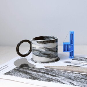 Puede incluir: Taza de cerámica hecha a mano con asa negra y diseño jaspeado gris y blanco. La taza está sobre una superficie impresa con varias herramientas de artesanía, incluyendo una herramienta de plástico azul y una cuchilla plateada.