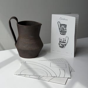 May include: A dark brown ceramic pitcher with a handle, next to a booklet with the word "Cántaros" and illustrations of pitchers. Several sheets of paper with architectural drawings are in the foreground. The items are on a white surface.