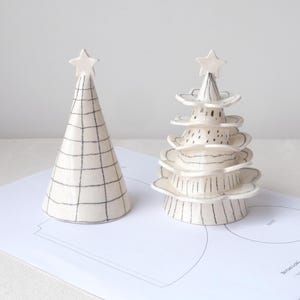 May include: Two ceramic Christmas trees. One is a cone shape with a grid pattern, topped with a star. The other is a tiered design with a star on top. Both are white with black detailing.