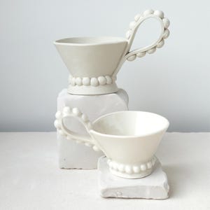 Puede incluir: Dos tazas de té de cerámica blanquecina con asas y bases únicas con cuentas. Las tazas tienen forma cónica y se apoyan sobre pedestales blancos texturizados. Las asas están adornadas con pequeñas cuentas redondas, añadiendo un toque decorativo.