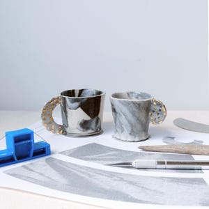 Puede incluir: Dos tazas de cerámica con diseños únicos. Una taza presenta un patrón veteado en blanco y negro, mientras que la otra tiene un efecto veteado gris y blanco. Ambas tazas tienen asas adornadas con detalles dorados. Las tazas están sobre una superficie blanca con útiles de arte.