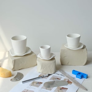 May include: Three white ceramic cups and saucers are displayed on beige blocks. Pottery tools, including a knife, a metal tool, and a blue plastic tool, are arranged on a white paper with text. A yellow sponge is also visible.