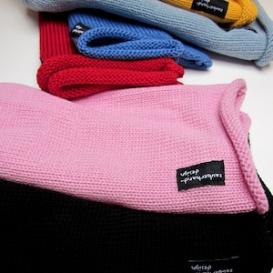 Könnte beinhalten: Eine Sammlung von gestrickten Mützen in verschiedenen Farben wie Pink, Schwarz, Rot, Blau und Gelb. Jede Mütze hat ein kleines schwarzes Stoffetikett mit dem weißen Text zauberhand-design. Der Text STYLE-fine-merino by zauberhanddesign ist sichtbar.