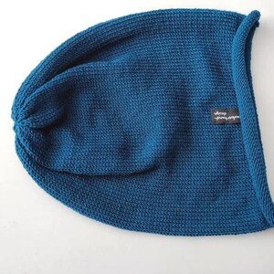 May include: A deep teal blue knit beanie hat featuring a rolled edge and a small black rectangular fabric tag on the side with the white text "uberhand design" printed on it. The hat has a consistent ribbed texture throughout the fabric.