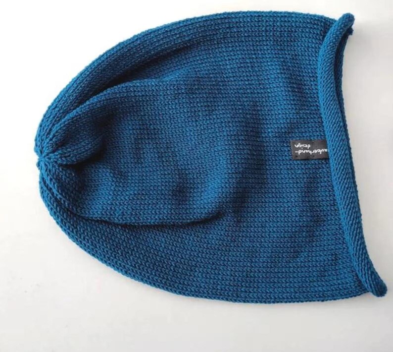 May include: A deep teal blue knit beanie hat featuring a rolled edge and a small black rectangular fabric tag on the side with the white text "uberhand design" printed on it. The hat has a consistent ribbed texture throughout the fabric.