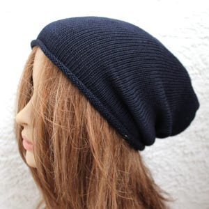 May include: A dark navy blue beanie hat with a fine knit texture and a simple rolled edge hem. The hat features a slouchy silhouette that sits loosely on the head, providing a relaxed fit with a smooth, uniform surface throughout the design.