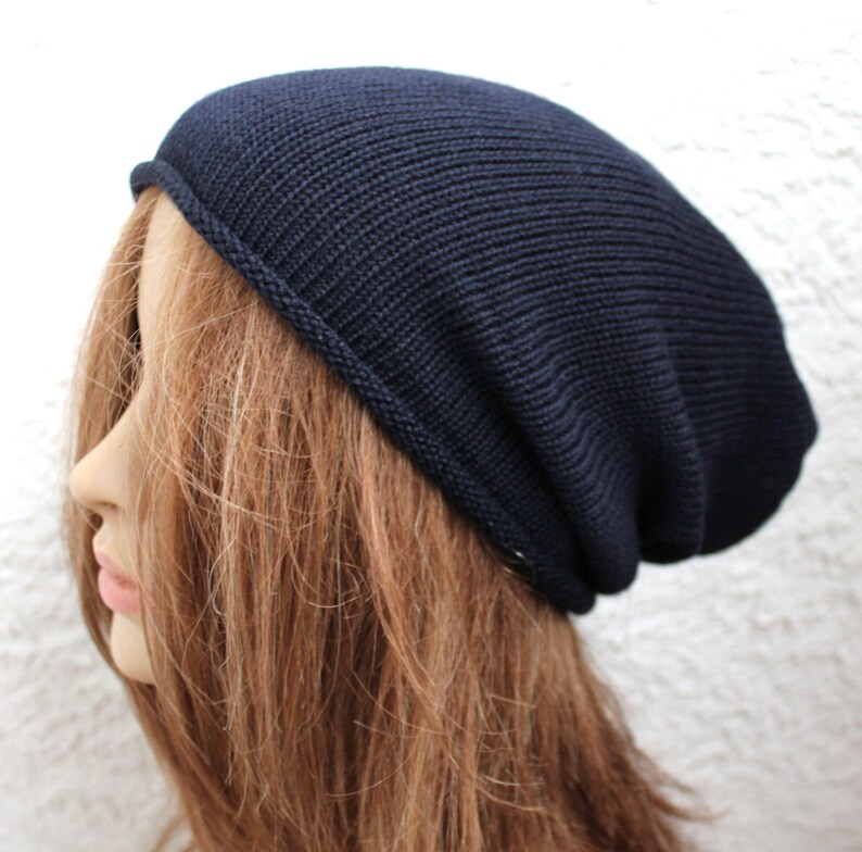 May include: A dark navy blue beanie hat with a fine knit texture and a simple rolled edge hem. The hat features a slouchy silhouette that sits loosely on the head, providing a relaxed fit with a smooth, uniform surface throughout the design.