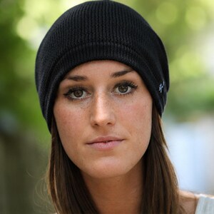 May include: A solid black knit beanie cap featuring a ribbed texture and a small white embroidered logo on the side. The hat has a rounded crown and a folded brim edge, constructed from a soft, flexible material that fits snugly around the head.