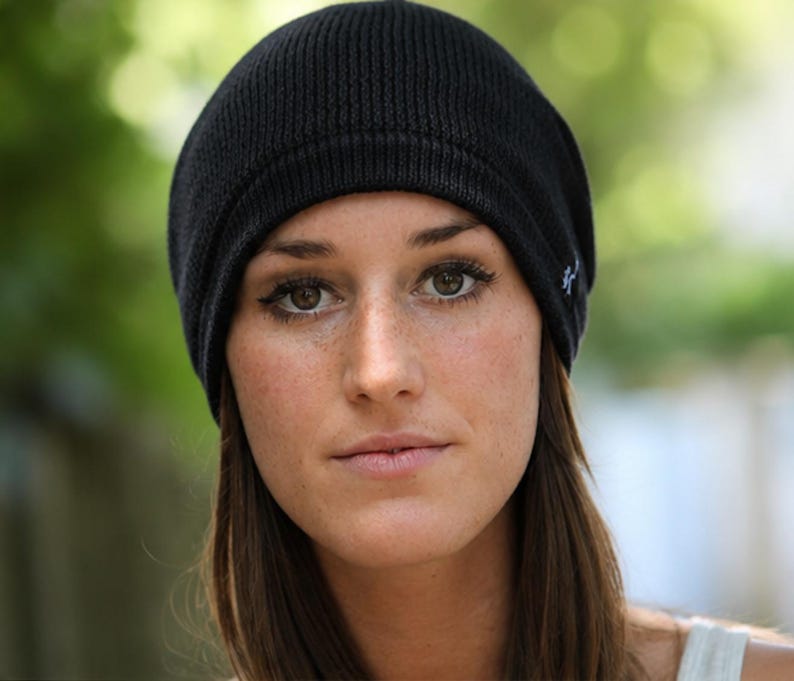 May include: A solid black knit beanie cap featuring a ribbed texture and a small white embroidered logo on the side. The hat has a rounded crown and a folded brim edge, constructed from a soft, flexible material that fits snugly around the head.