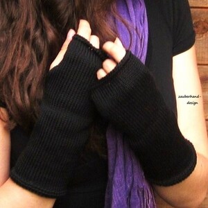 May include: A pair of black fingerless gloves with a ribbed knit texture. The gloves are worn by a person with long brown hair and a purple scarf.