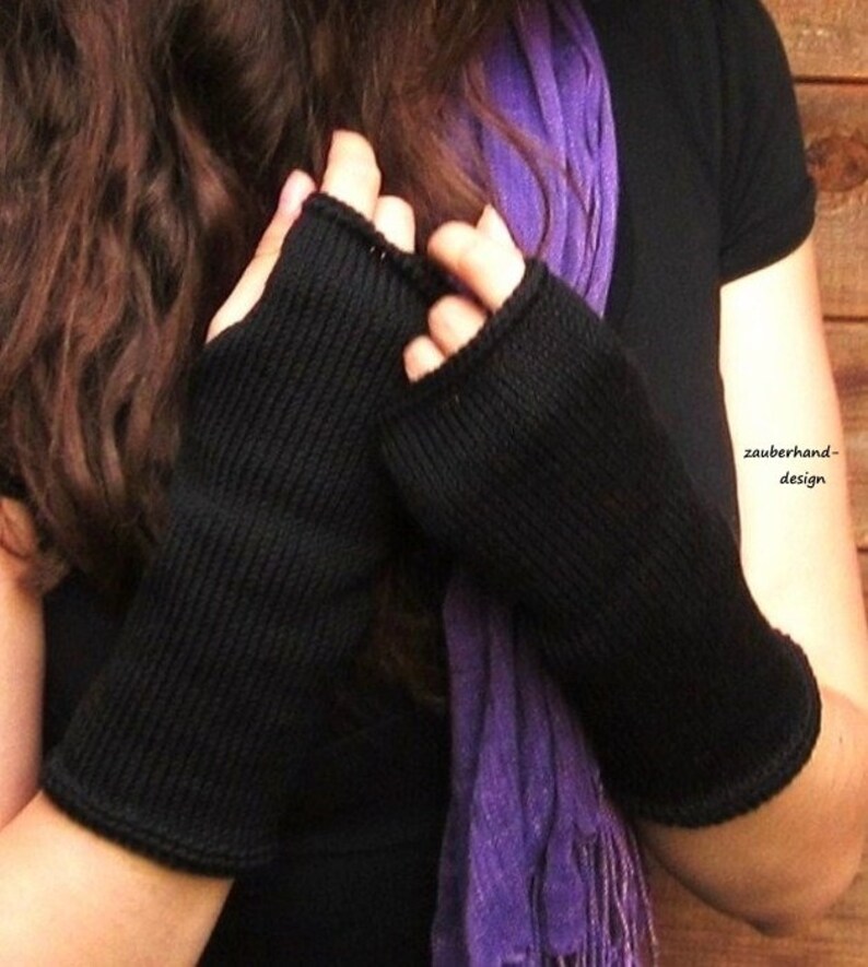 May include: A pair of black fingerless gloves with a ribbed knit texture. The gloves are worn by a person with long brown hair and a purple scarf.