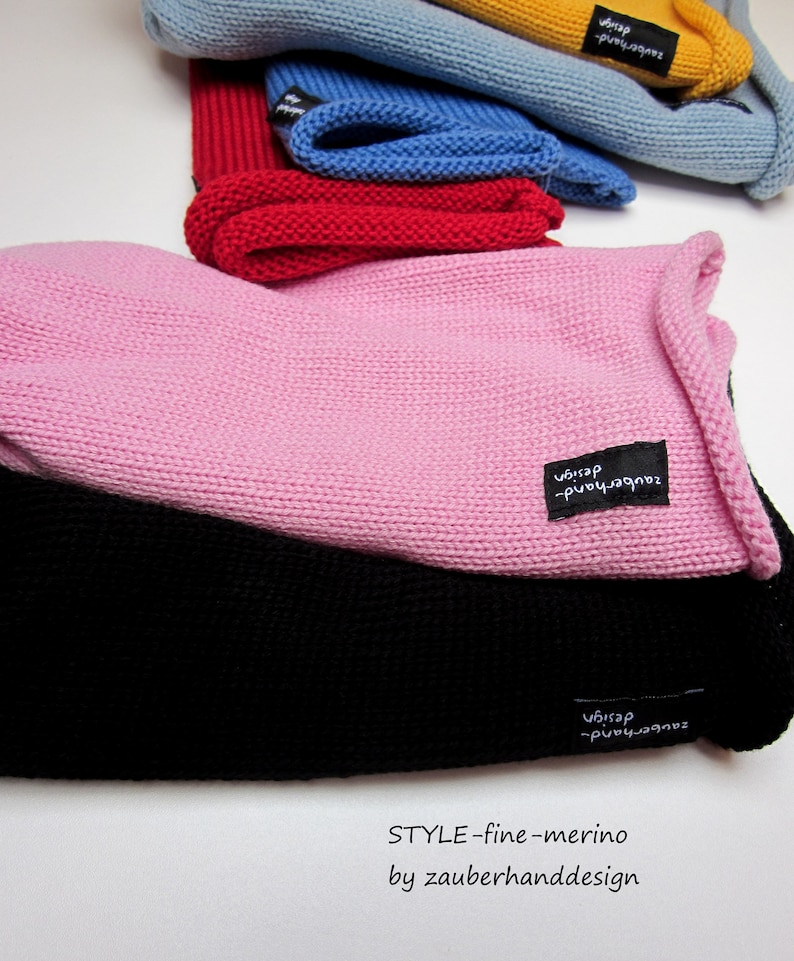 Könnte beinhalten: Eine Sammlung von gestrickten Mützen in verschiedenen Farben wie Pink, Schwarz, Rot, Blau und Gelb. Jede Mütze hat ein kleines schwarzes Stoffetikett mit dem weißen Text zauberhand-design. Der Text STYLE-fine-merino by zauberhanddesign ist sichtbar.