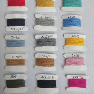 May include: Fifteen spools of yarn in various colors including red, mustard, turquoise, midnight blue, light gray, azure, taupe, dark gray, yellow, camel, fudge, rose, stone, black, and fuchsia. Each spool has a handwritten label above it.