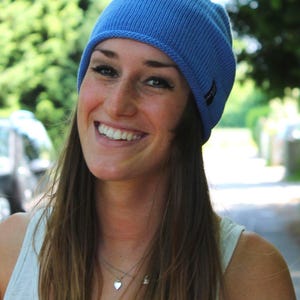 May include: A light blue ribbed knit beanie cap featuring a small dark rectangular brand tag on the side. The hat is constructed with a vertical ribbed texture and a rolled edge, providing a snug fit around the head with a consistent solid color.