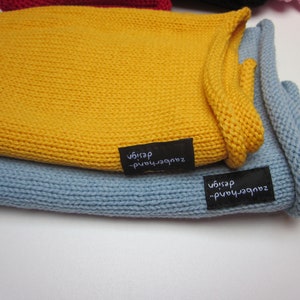 May include: A mustard yellow knitted fabric piece layered over a light blue knitted fabric piece. Each item features a small black rectangular fabric tag sewn onto the edge with the white embroidered text zauberhand-design visible on both.