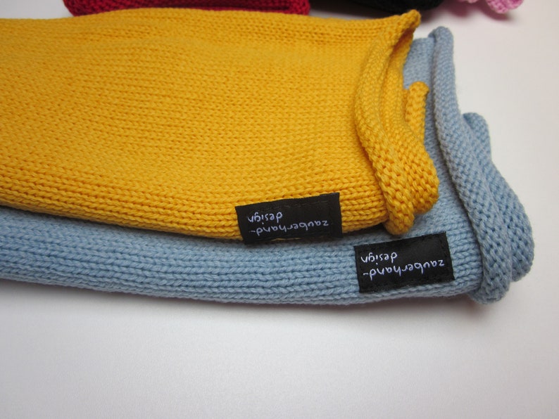 May include: A mustard yellow knitted fabric piece layered over a light blue knitted fabric piece. Each item features a small black rectangular fabric tag sewn onto the edge with the white embroidered text zauberhand-design visible on both.