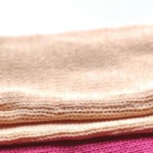 May include: Two knitted blankets, one peach and one pink, are folded and stacked on top of each other. The peach blanket has a black label with the text "wrap yourself in warmth" on it. The pink blanket has a black label with the text "wrap yourself in warmth" on it.