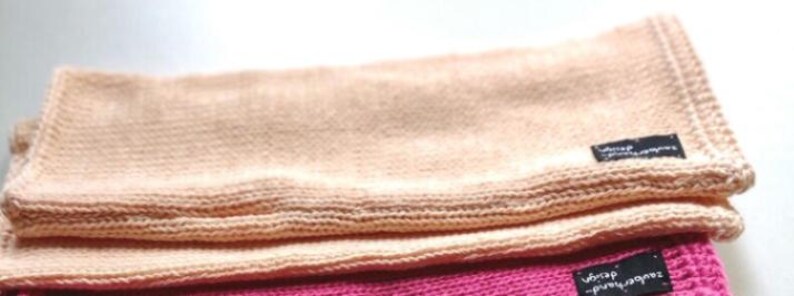 May include: Two knitted blankets, one peach and one pink, are folded and stacked on top of each other. The peach blanket has a black label with the text "wrap yourself in warmth" on it. The pink blanket has a black label with the text "wrap yourself in warmth" on it.