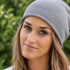May include: A light gray knit beanie with a smooth, fine-gauge texture and a simple rolled edge. The hat features a rounded crown design that fits closely to the head, crafted from a soft, uniform knit fabric in a neutral, solid light gray color.
