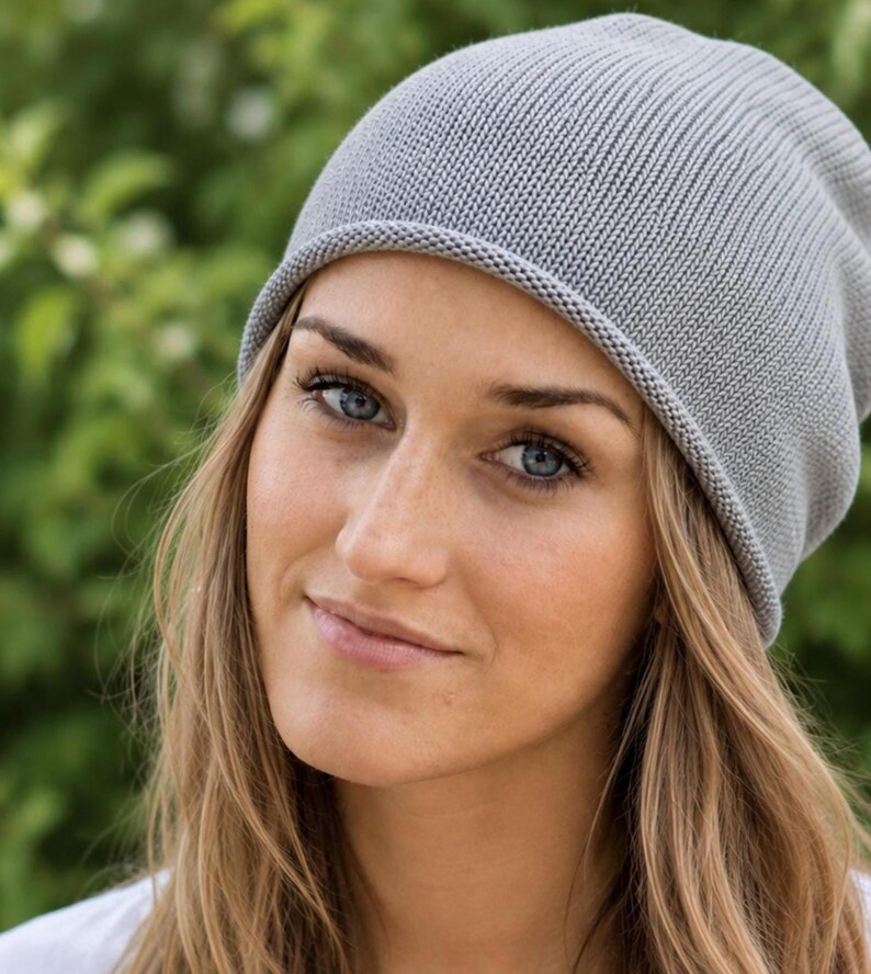May include: A light gray knit beanie with a smooth, fine-gauge texture and a simple rolled edge. The hat features a rounded crown design that fits closely to the head, crafted from a soft, uniform knit fabric in a neutral, solid light gray color.